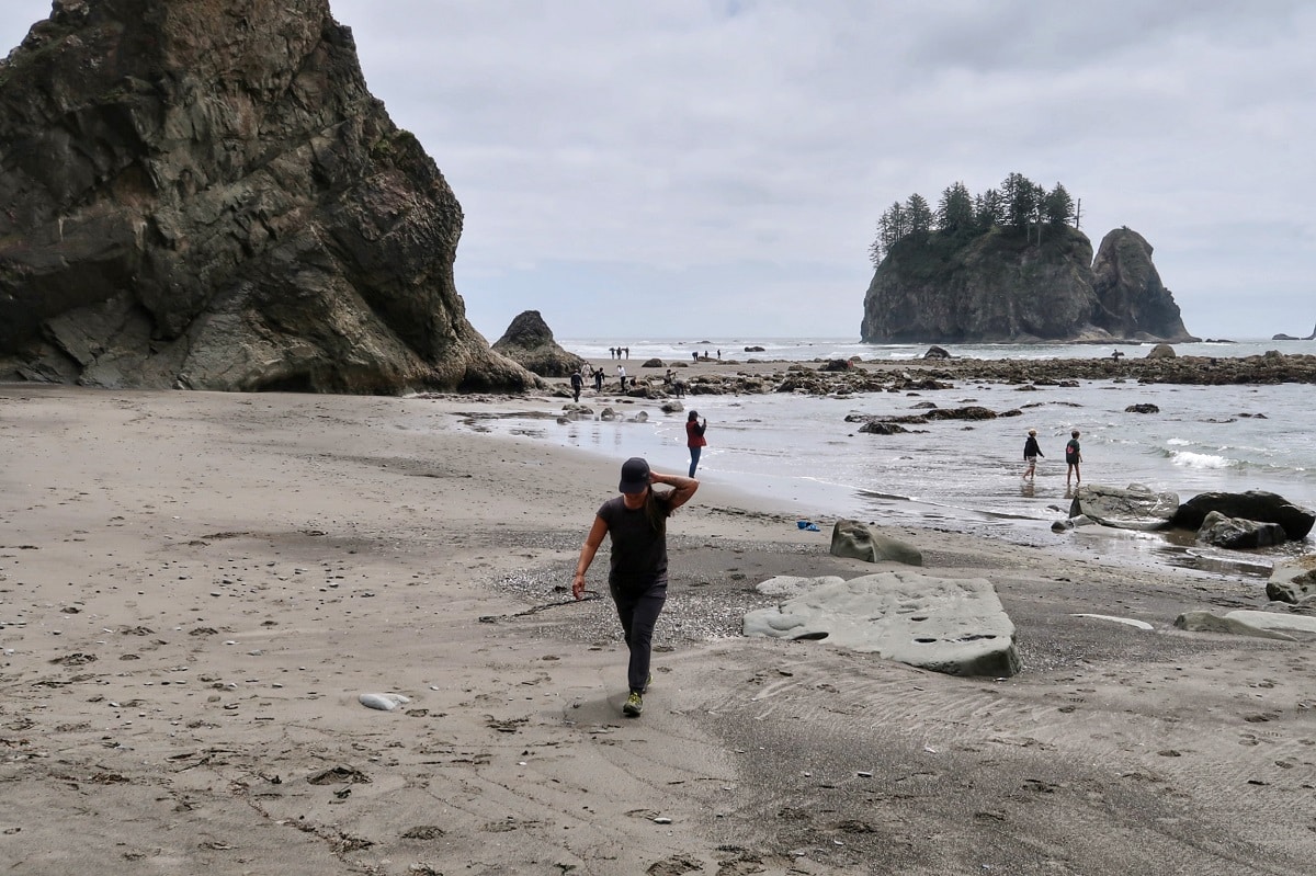 woman walks toward the person taking her photo on a sandy beach with big rocks and the ocean behind her