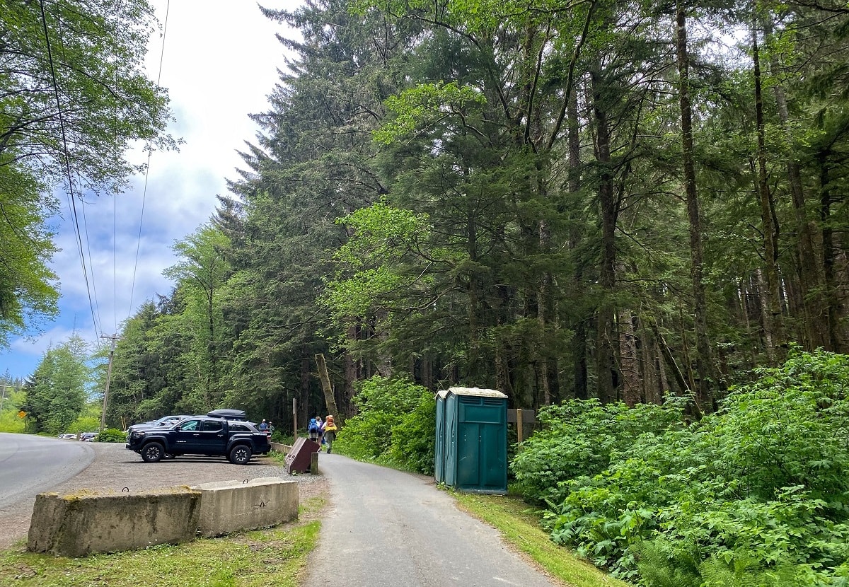 walking path to a trailhead next to port-o-potties and a parking area, surrounded by forest