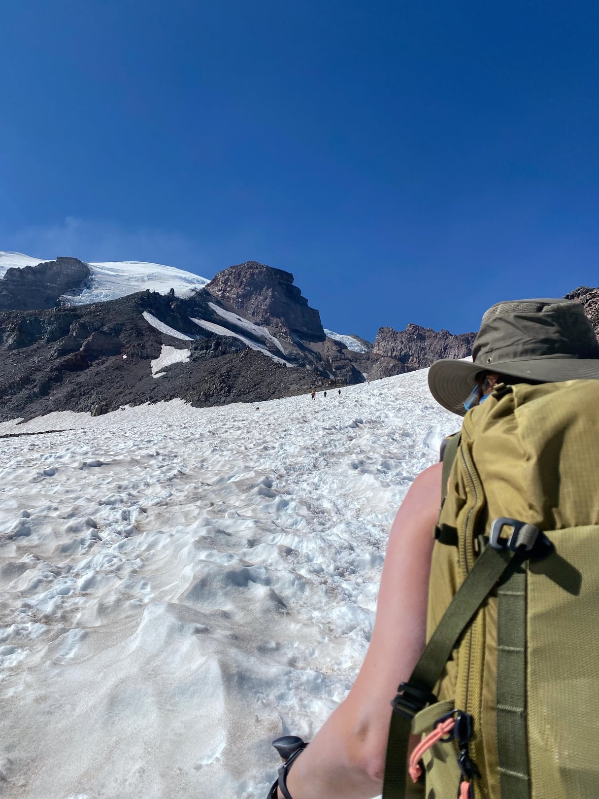 Over-the-shoulder view of a hiker on the Camp Muir trail wearing a green backpack on the snow-covered slopes of Mount Rainier under a clear blue sky. 