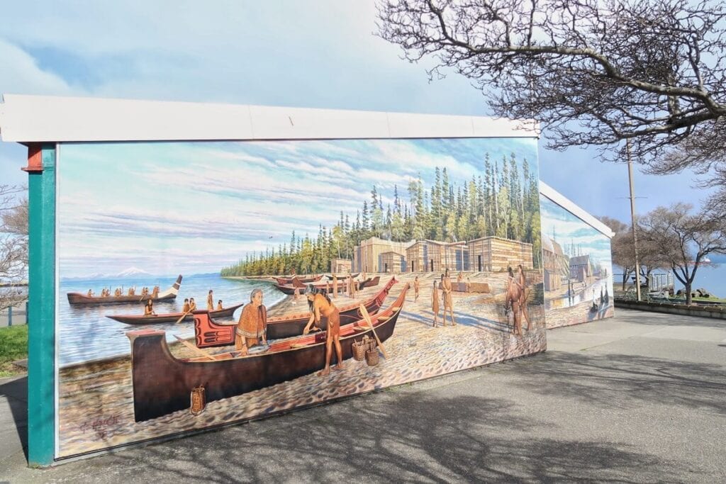 Vivid mural depicting historic canoe scene on a building's side, under a bare tree against a bright sky.