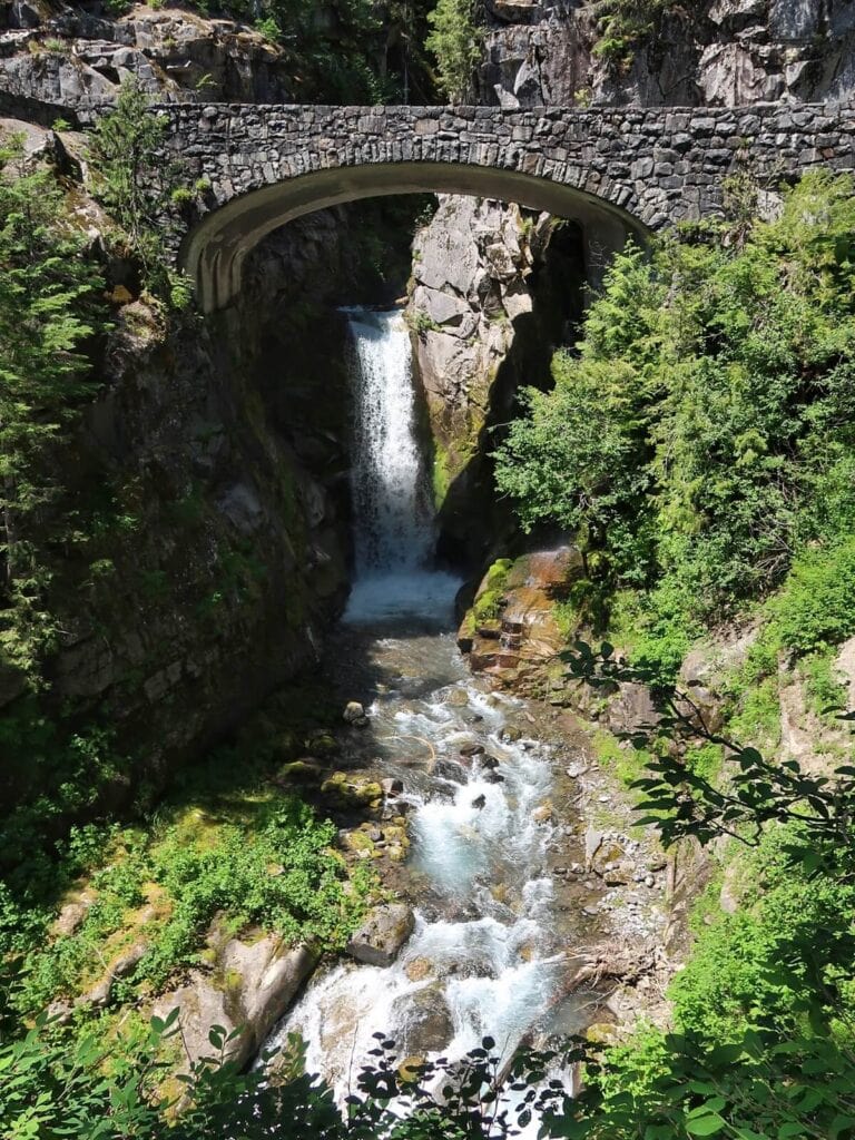 Christine Falls cascading gently beneath a picturesque stone bridge. The waterfall is surrounded by lush greenery and moss-covered rocks, with the flowing water creating a serene and tranquil scene. 