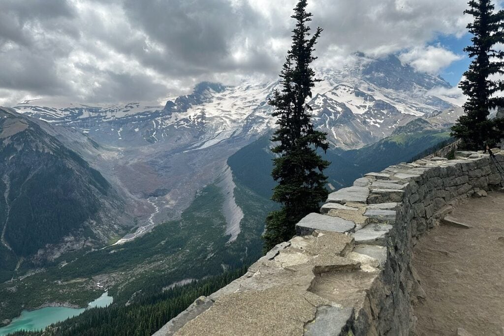 A breathtaking mountain view from Glacier Overlook on the Sunrise Rim Trail, with a stone wall in the foreground framing the dramatic, rugged scene. Below, a glacier-fed valley and a turquoise alpine lake are surrounded by dense evergreen forests.