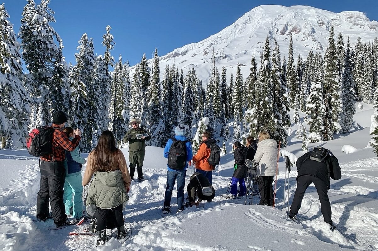 A group of people on a ranger-guided snowshoe walk at Paradise, one of the best Mt Rainier activities in winter, surrounded by snow-covered evergreen trees. The ranger, dressed in green, speaks to the group against the backdrop of the Mountain’s towering, snow-covered slopes. 