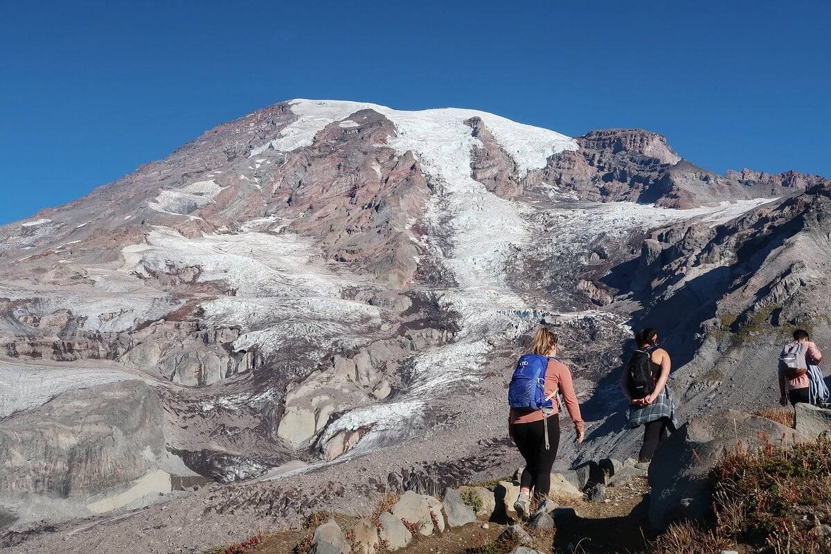 Hikers traverse a rugged trail with the Nisqually Glacier and the massive, rocky slopes of the Mountain dominating the background. The glacier’s icy surface contrasts with the exposed brown and gray mountain terrain.