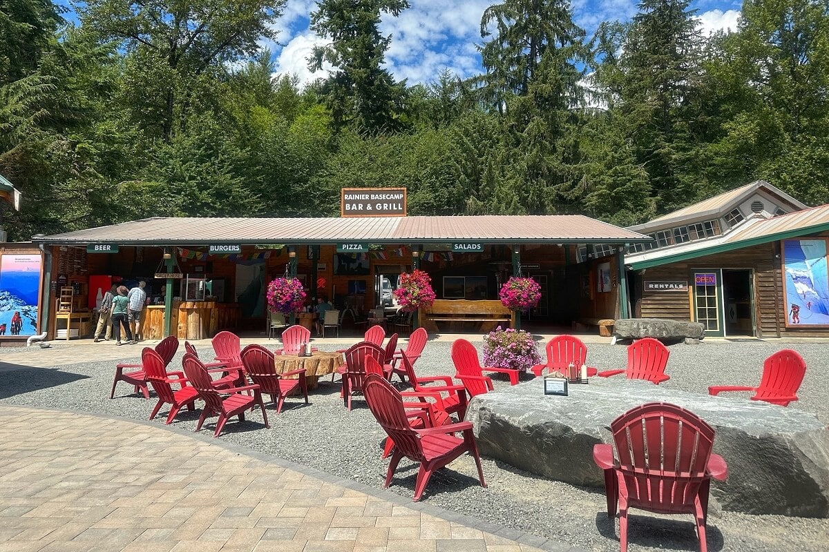 The Rainier BaseCamp Bar and Grill, nestled among lush evergreen trees, features a rustic outdoor seating area with red Adirondack chairs arranged around a fire pit. It offers food and drink options like beer, burgers, pizza, and salads, and colorful hanging flower baskets add charm to the scene. This inviting setting provides a cozy spot for visitors to relax after exploring and hiking.
