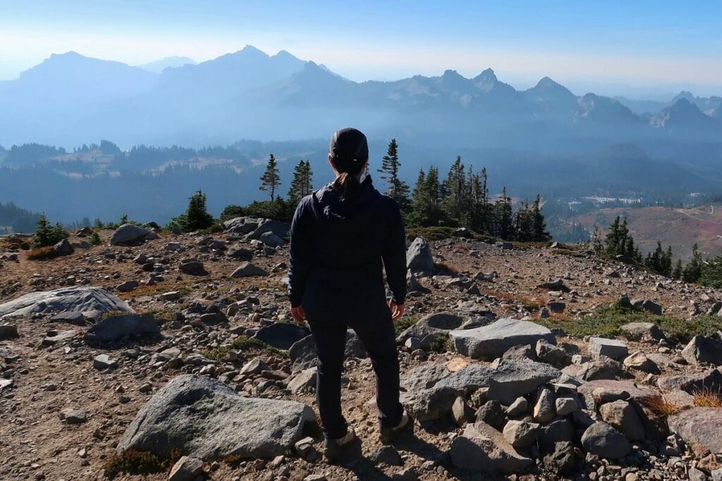 A person stands on rocky terrain on one of the best hikes in Mount Rainier, gazing out at a breathtaking view of distant mountain peaks partially obscured by a soft blue haze. The foreground features scattered boulders and alpine vegetation, with tall evergreen trees framing the midground. 