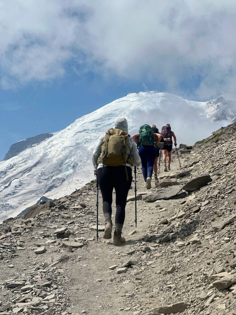A group of hikers equipped with backpacks and trekking poles ascend the rocky Burroughs Mountain trail toward Rainier’s snow-covered slopes. The path is steep and rugged, surrounded by barren terrain, with the glacier looming under a partly cloudy sky. 