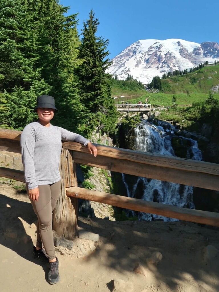 A smiling person leans against a wooden railing from the iconic viewpoint overlooking Myrtle Falls. The waterfall cascades down a lush green hillside, with Rainier's snow-capped peak towering in the background under a clear blue sky.