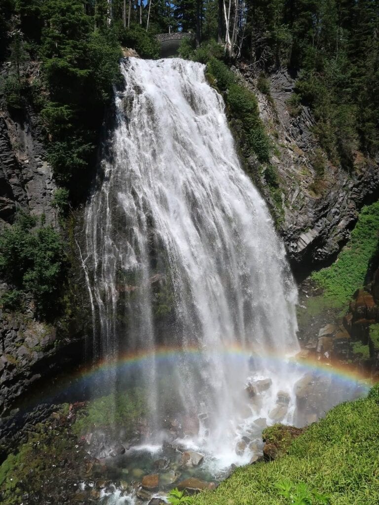 A magnificent Mt Rainier attraction—Narada Falls cascading down a rocky cliff surrounded by lush greenery and tall evergreen trees. At the base of the falls, a vibrant rainbow arcs through the mist, adding a colorful touch to the scene. 