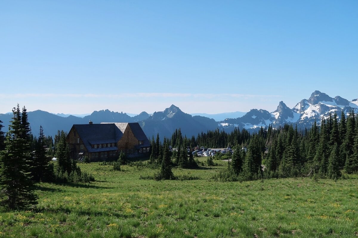 A scenic view from the Sunrise Nature Trail, featuring a rustic lodge nestled among green meadows and surrounded by evergreen trees. Snow-capped mountain peaks rise in the background under a clear blue sky, while a parking area is visible nearby. 
