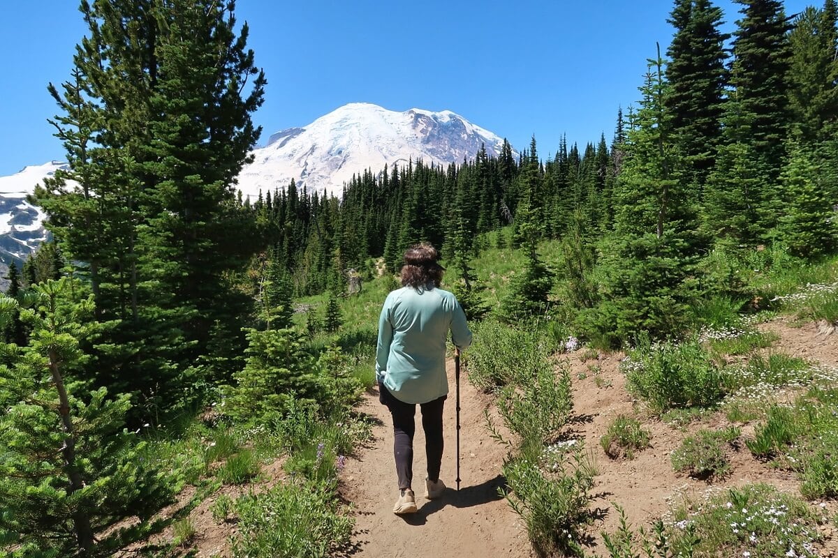 A hiker wearing a light blue long-sleeve shirt and carrying a trekking pole walks along a dirt trail surrounded by lush green evergreens. Rainier’s snow-capped peak towers in the background under a bright blue sky. 