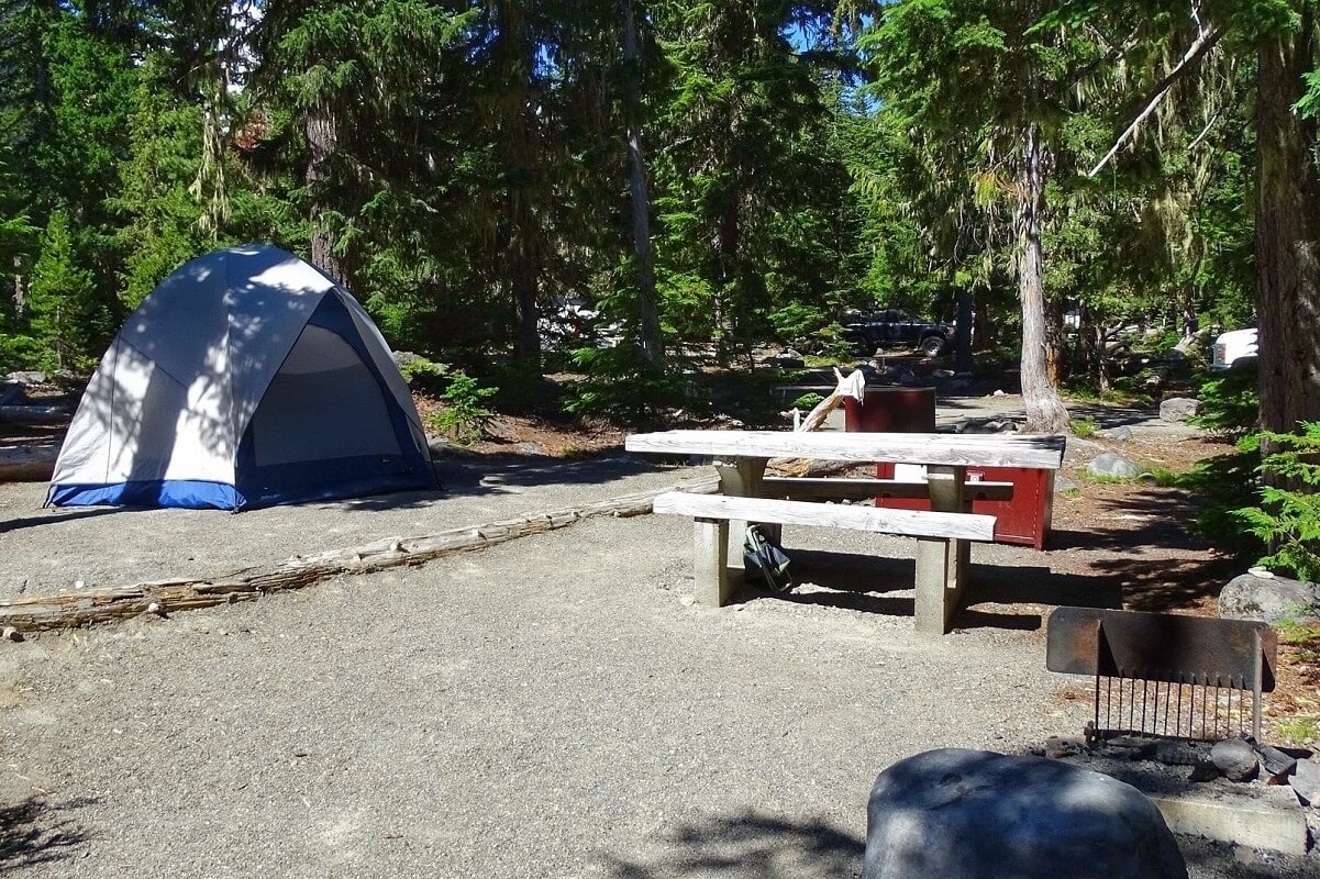 A campsite in the White River Campground at Mount Rainier National Park, featuring a pitched blue and white tent surrounded by tall evergreen trees. The site includes a wooden picnic table, a fire pit, and a food storage box for safety. 