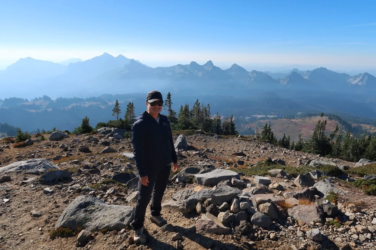 A smiling woman on the Skyline Loop Trail at Mount Rainier in a dark jacket, sunglasses, and a cap. Behind her, a stunning view of jagged mountain peaks under a clear blue sky fades into the horizon. The scene captures the expansive and serene beauty of a high-altitude landscape.