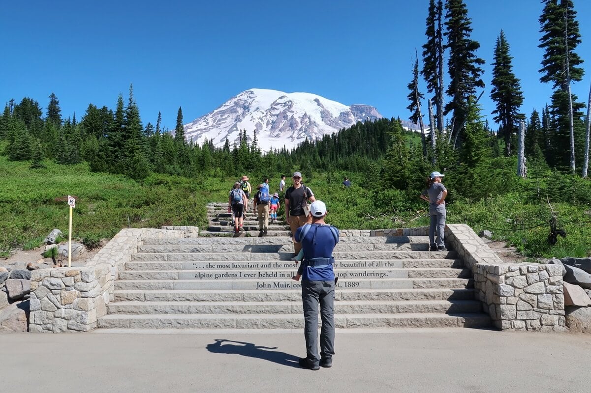 Visitors climb the iconic Muir Steps at Mount Rainier National Park, with the snow-capped peak of the Mountain towering in the background under a clear blue sky. The stone steps are inscribed with a quote from John Muir: "…the most luxuriant and the most extravagantly beautiful of all the alpine gardens I ever beheld in all my mountain-top wanderings." Lush greenery and tall evergreens frame the scene.