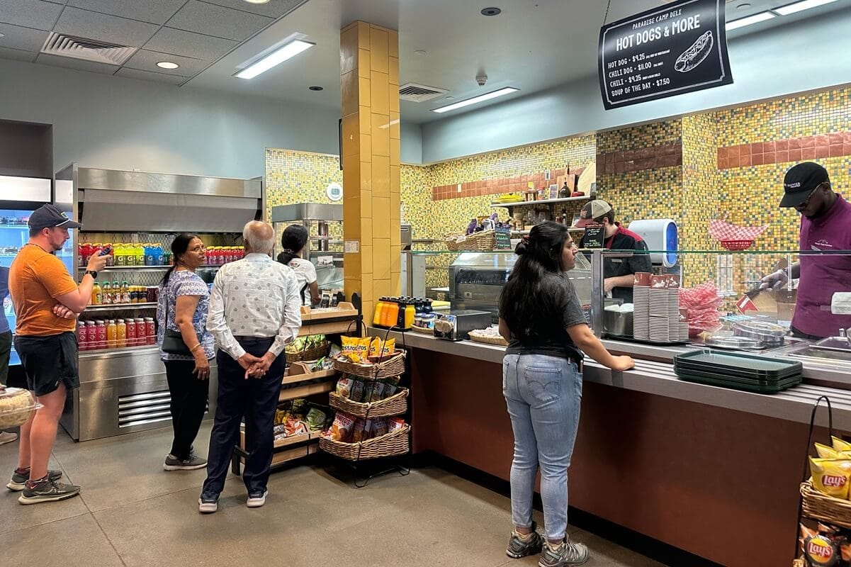 Customers line up at the Paradise Camp Deli, a casual eatery with a menu offering hot dogs, chili, and soup of the day, displayed on a hanging sign above the counter. Staff members behind the counter prepare orders, while visitors browse or wait for their food in the relaxed atmosphere.