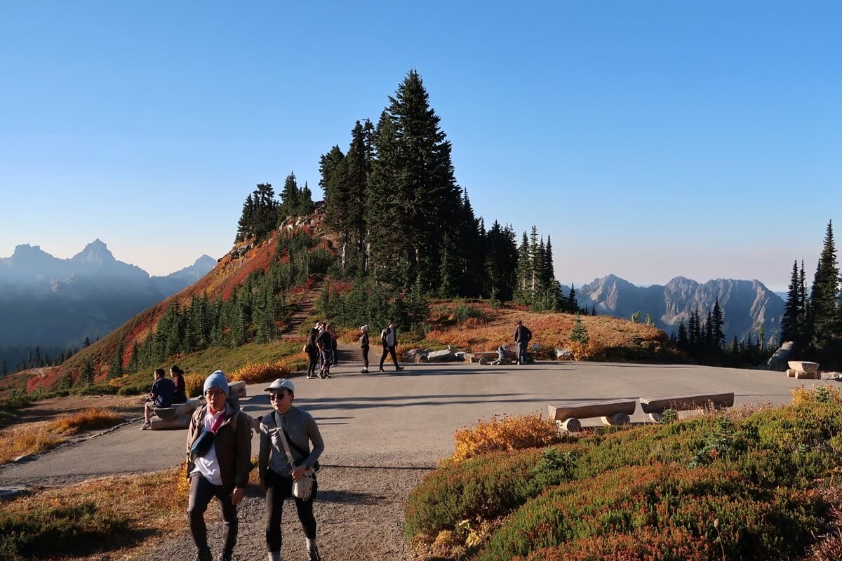 Hikers walking along a paved path on the Skyline Loop Trail, surrounded by vibrant fall foliage. The scene features colorful autumn vegetation, towering pine trees, and a backdrop of rugged mountain peaks under a clear blue sky. 