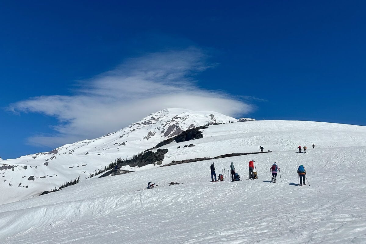 People hiking on a snow-covered slope under a clear blue sky, with a towering snow-capped mountain in the background. The scene shows individuals carrying trekking poles, some resting, as they navigate the bright and expansive snowy terrain.