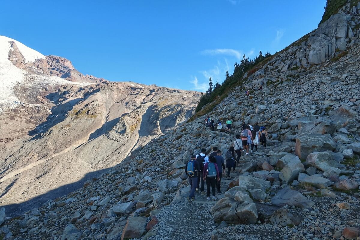 A group of hikers ascends the rocky Skyline Trail toward Panorama Point, with the rugged slopes of Rainier visible to the left under a clear blue sky. The narrow trail weaves through a steep, boulder-strewn landscape, showcasing the raw and dramatic alpine terrain. 