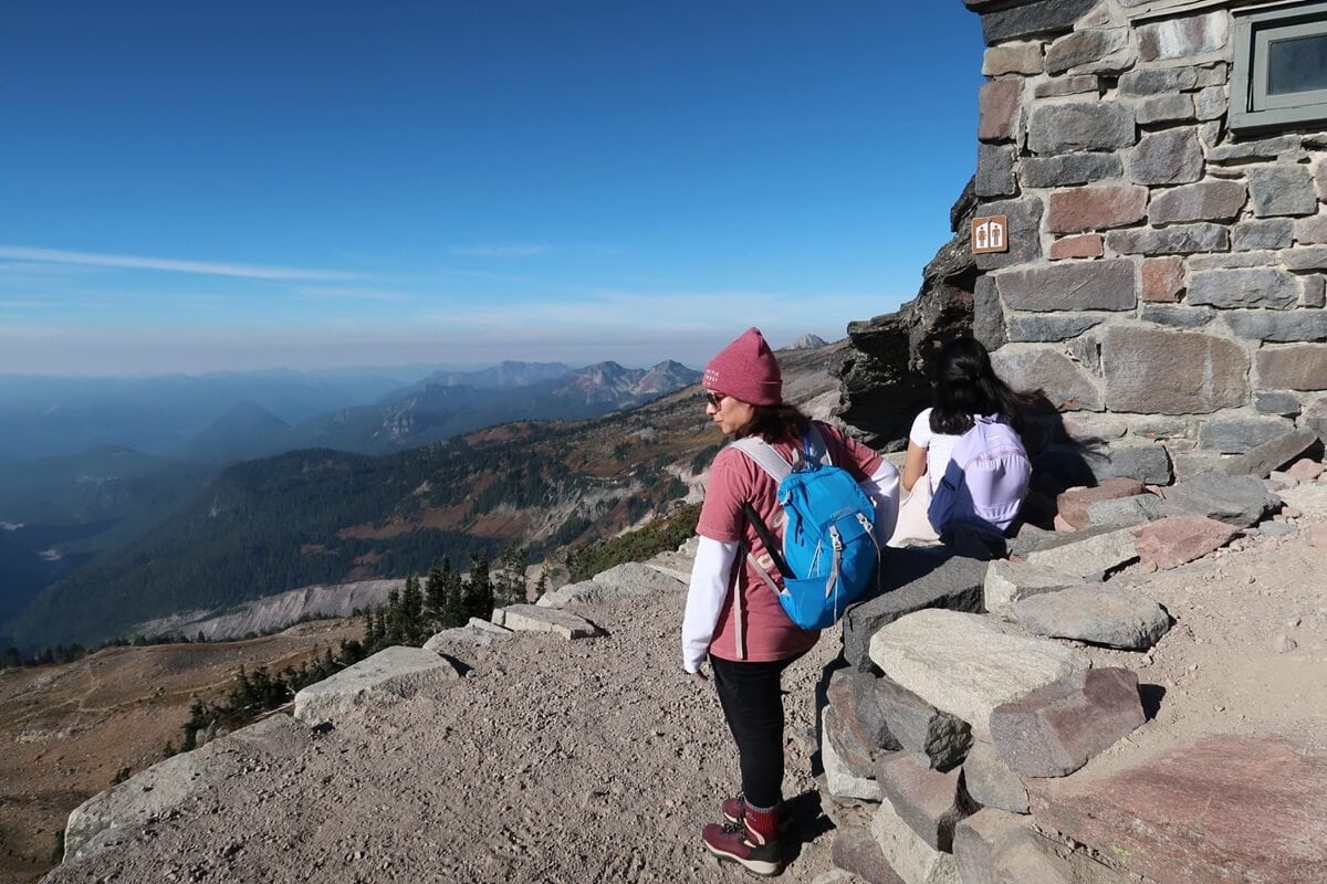 Two hikers resting near a stone structure bathroom at Panorama Point, with one wearing a blue backpack and a pink beanie, standing and looking out at the sweeping mountain and valley views. The other hiker sits on the rocky ledge, facing away, under a bright blue sky with distant mountain ranges in view.