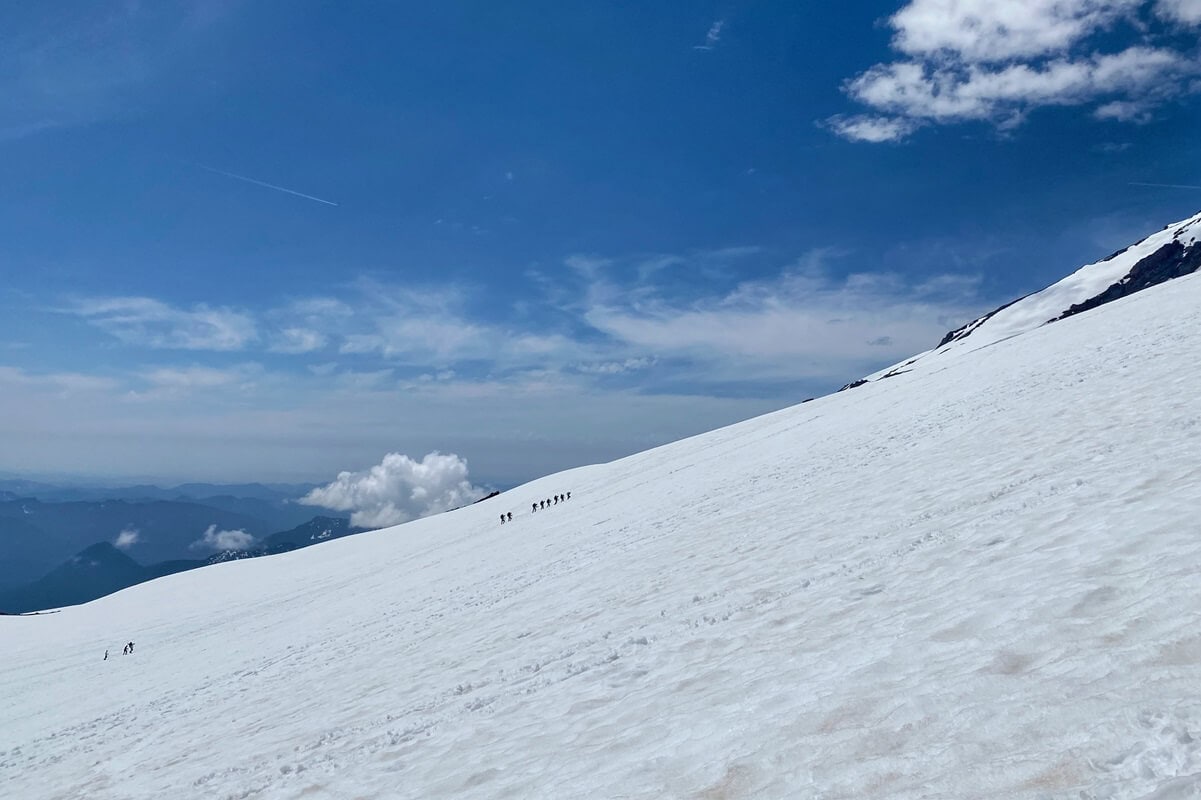 A line of climbers ascends the expansive Muir Snowfield on Mount Rainier, surrounded by an endless stretch of snow under a bright blue sky with scattered clouds. The image captures the vastness and challenge of alpine climbing in a pristine and remote environment.
