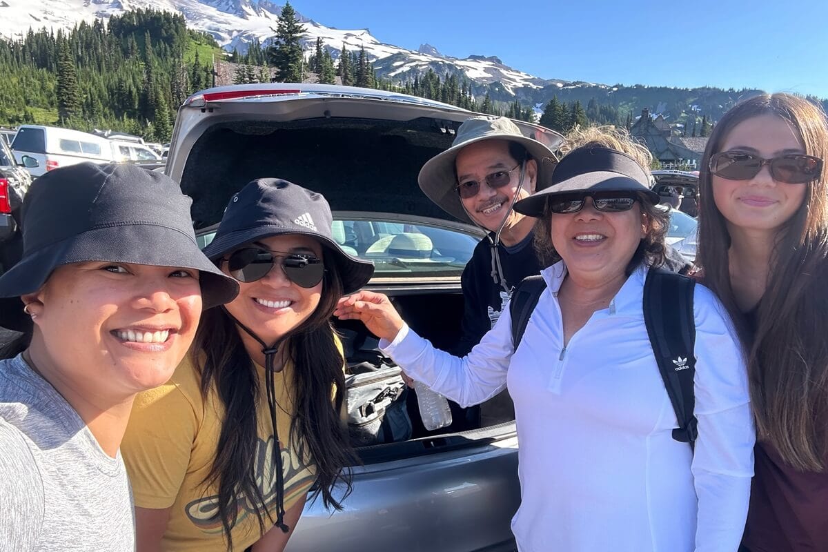A family smiling and posing in front of an open car trunk at the main Paradise parking lot, with a scenic backdrop of snow-capped mountains, evergreen trees, and clear blue skies. Everyone is wearing casual outdoor clothing, sun hats, and sunglasses, prepared for a day in nature.