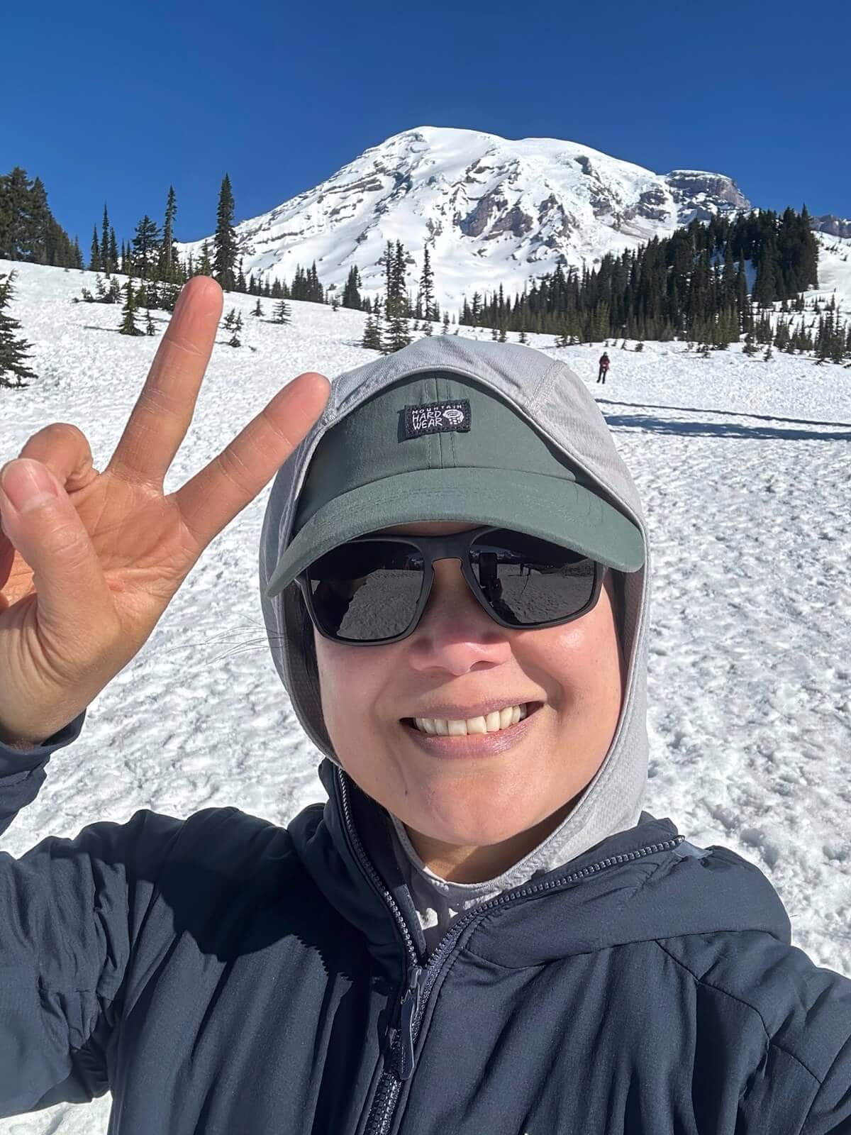 A smiling woman taking a selfie on a snowy trail, wearing sunglasses, a hooded jacket, and a hat, while holding up a peace sign. In the background, a snow-covered mountain and evergreen trees are visible under a bright blue sky.