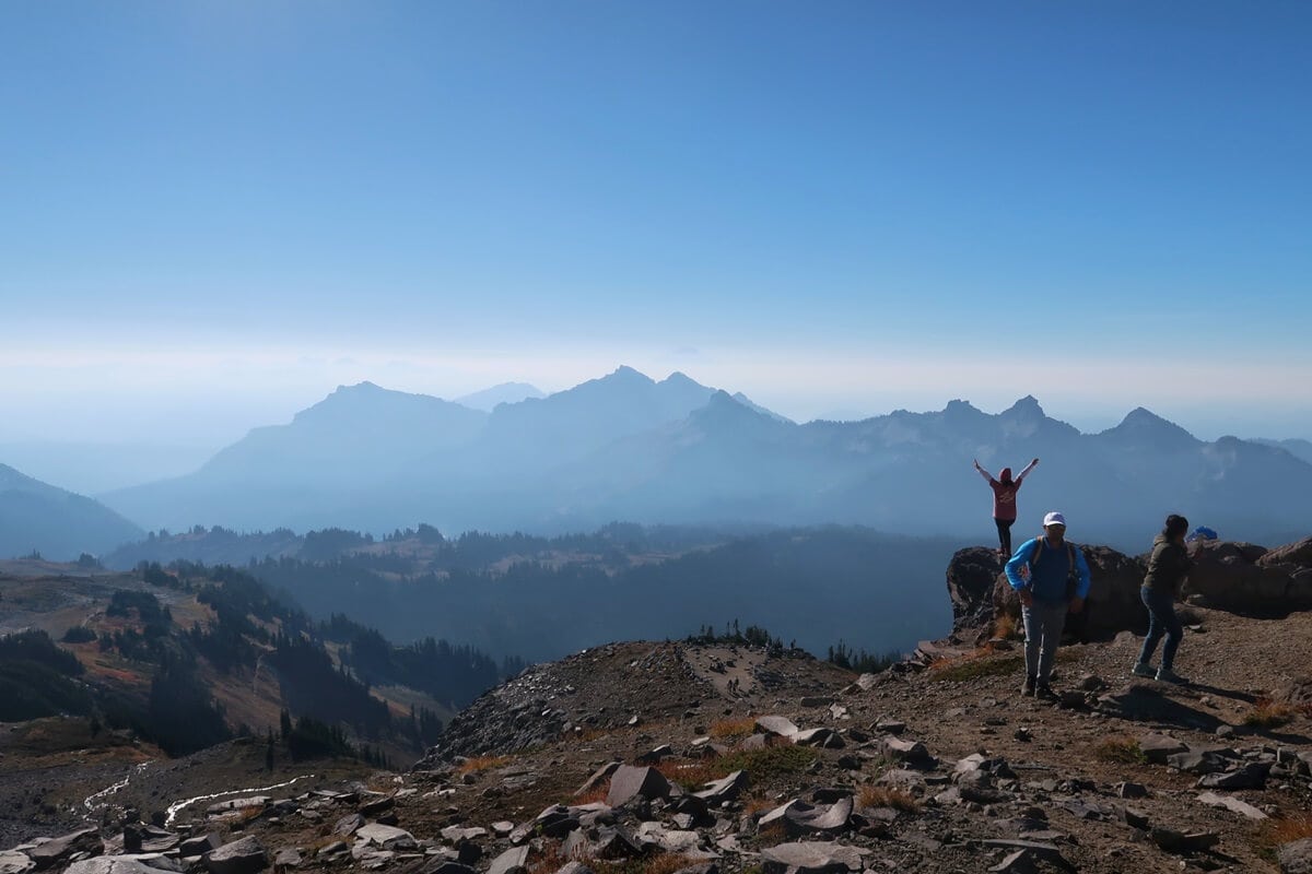 A breathtaking view from Panorama Point features layers of hazy mountain ridges stretching into the horizon under a clear blue sky. In the foreground, a hiker stands triumphantly on a rocky outcrop with arms raised, while others explore the rugged terrain nearby. The scene captures the vastness and beauty of the alpine landscape, inviting a sense of awe and adventure.