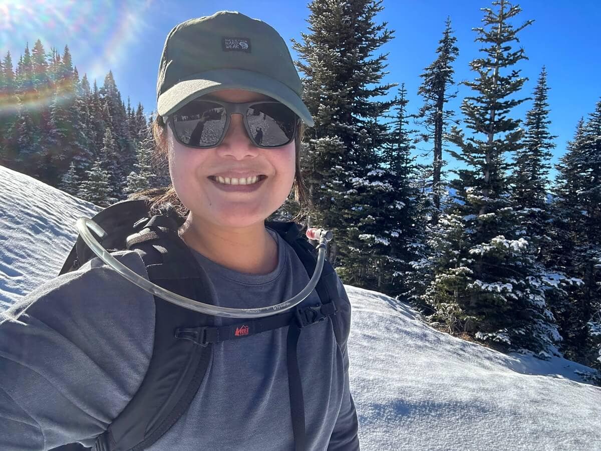 Smiling woman takes a selfie at Hurricane Ridge in January under a clear blue sky. She's wearing sunglasses, a dark green cap, a long-sleeve base layer, and a hydration pack, with sun flare visible in the upper left. 