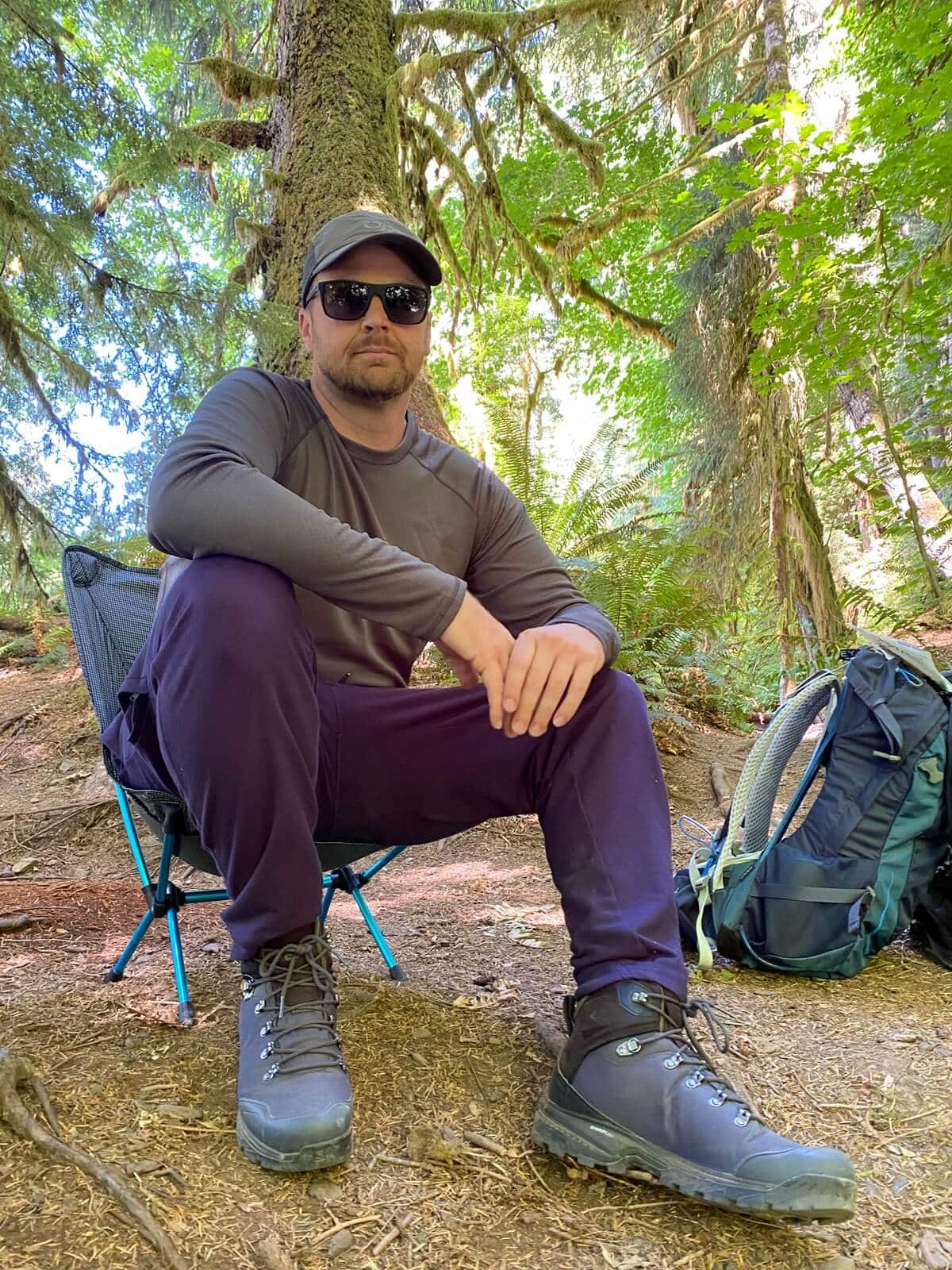Man in the Hoh Rain Forest wearing sunglasses, a long-sleeve hiking shirt, dark hiking pants, and sturdy lace-up boots sits on a low hiking chair. A hiking backpack rests nearby, and tall trees with moss-draped branches surround the rest site.