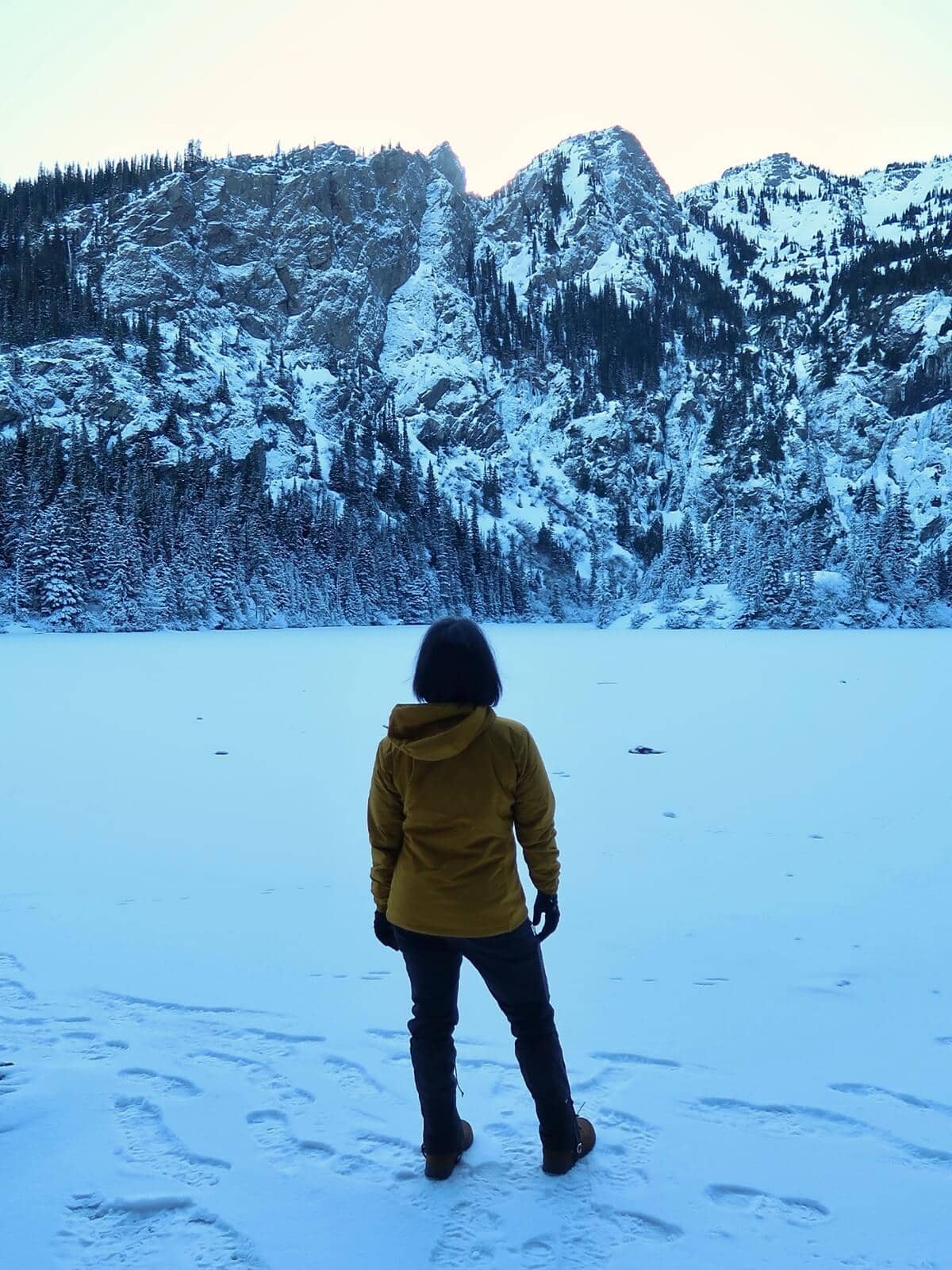 A woman dressed in winter hiking gear, including insulated boots, gloves, and a mustard-yellow jacket, stands on a snow-covered lake in Olympic National Park, facing a rugged, snow-draped mountain ridge.