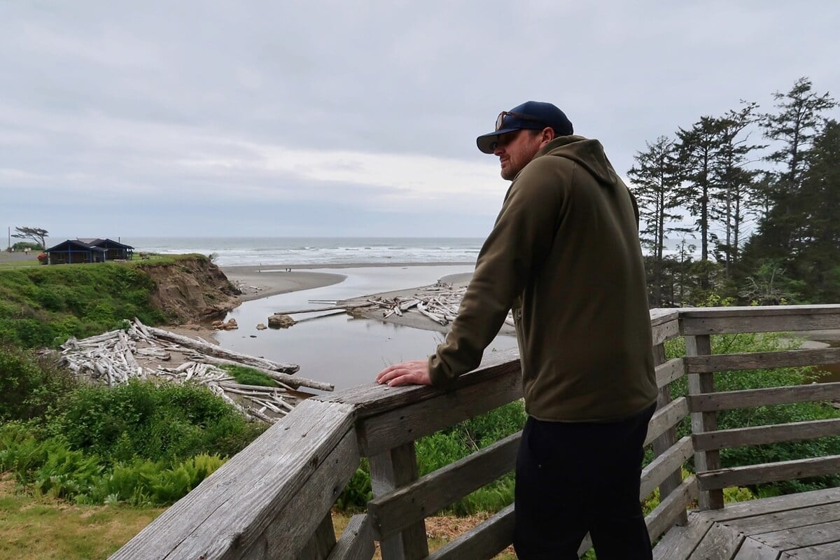 A person stands on a rustic wooden viewing platform at Kalaloch Lodge, overlooking a serene beach scattered with driftwood logs. The overcast sky casts a soft light over the rugged coastline, where the ocean meets a sandy shore lined with lush greenery and scattered logs.