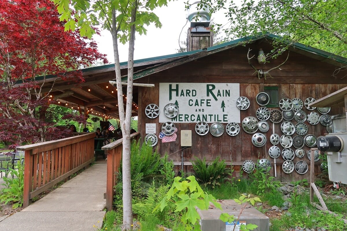 A rustic wooden building labeled "Hard Rain Café and Mercantile," decorated with dozens of metal hubcaps arranged on the exterior wall. The café features a covered patio strung with lights, surrounded by lush green plants and vibrant red and green trees.