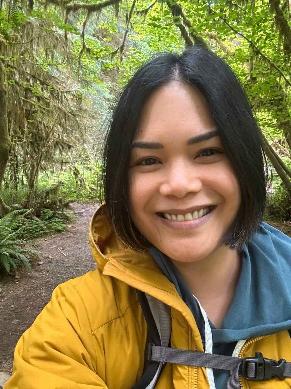 A smiling hiker on a lush, forested Hoh Rain Forest trail in a bright yellow rain jacket takes a selfie while walking. The background is filled with moss-draped trees, ferns, and vibrant greenery, capturing the misty, enchanting beauty of the temperate rainforest.
