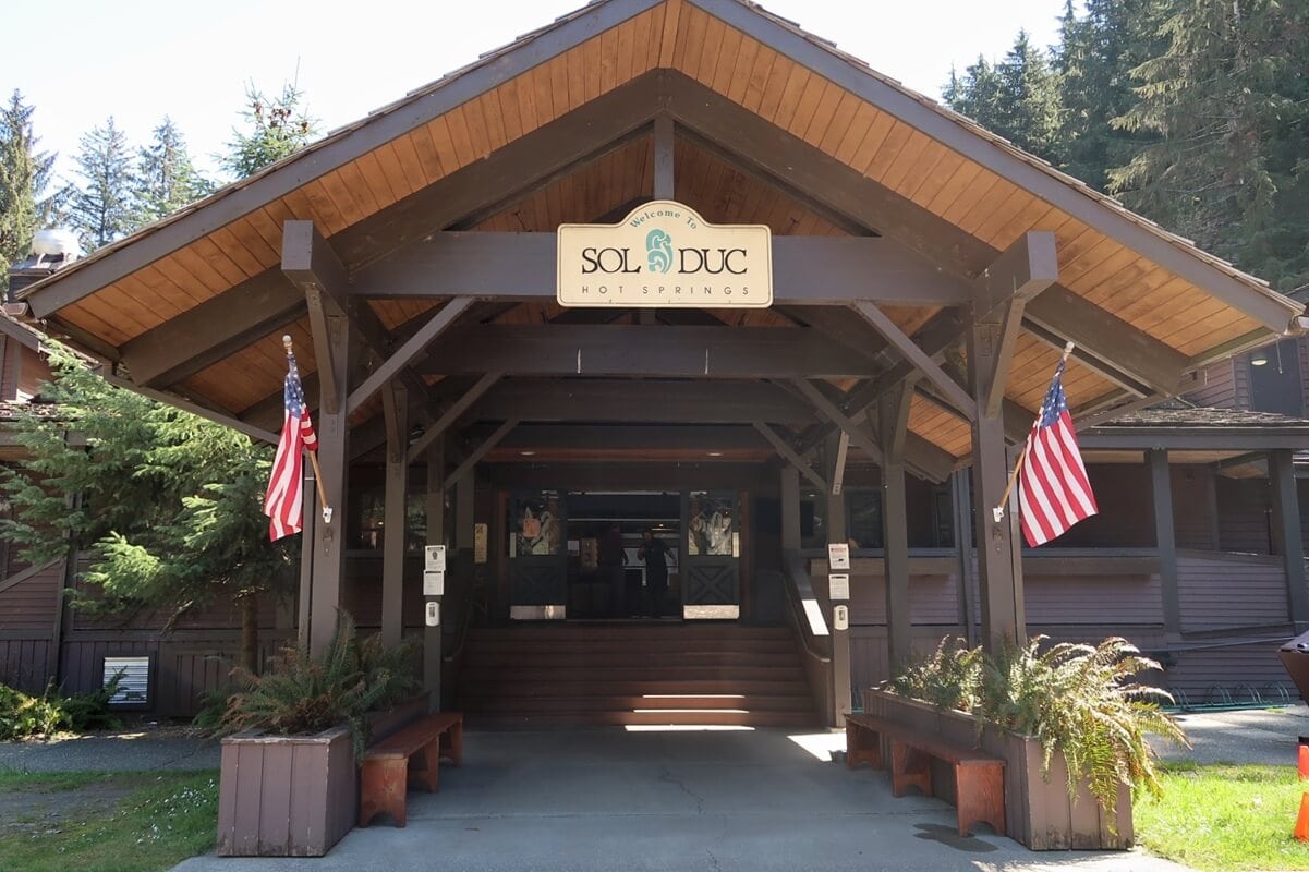 The entrance to Sol Duc Hot Springs Resort features a rustic wooden canopy with a sign reading “Welcome to Sol Duc Hot Springs,” flanked by two American flags. Surrounded by evergreens and tucked into the valley, the resort offers a cozy gateway to the region’s natural beauty and relaxation.