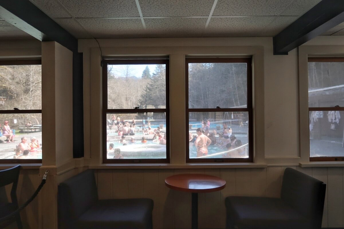 View through a window at Sol Duc Hot Springs, showing people relaxing in the outdoor thermal pools surrounded by forested hills and crisp mountain air. The warm, mineral-rich water draws visitors seeking a soothing soak.