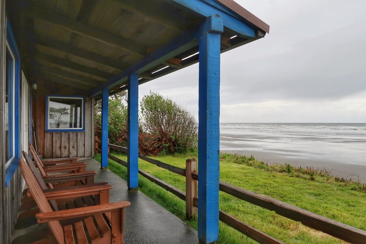 Covered porch of a Kalaloch Lodge cabin, with wooden chairs facing a quiet, overcast beach framed by bright blue beams and rustic wood paneling. The lawn leads to the shoreline, offering peaceful ocean views and a cozy spot to relax by the sea.