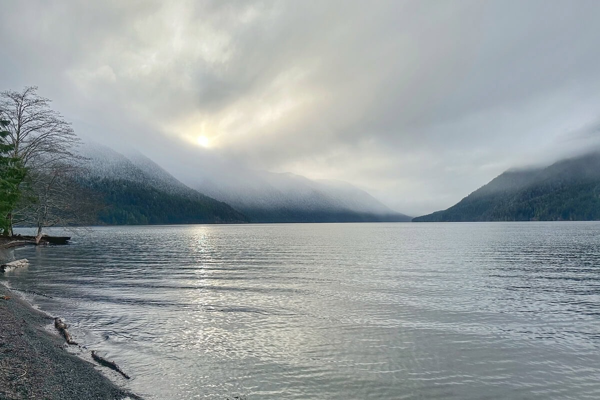 Lake Crescent in Olympic National Park in winter with misty clouds hanging low over snow-dusted mountains and calm, reflective waters. The sun peeks faintly through the overcast sky, casting a soft glow across the lake’s surface near a quiet, tree-lined shoreline.
