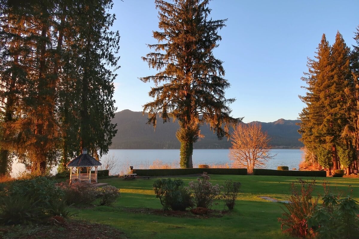 Lakeside garden view at Lake Quinault Lodge during golden hour, with sunlight casting a warm glow on tall evergreens and a bare deciduous tree. A small white gazebo sits near the water’s edge, framed by manicured lawns and mountain ridges in the background.