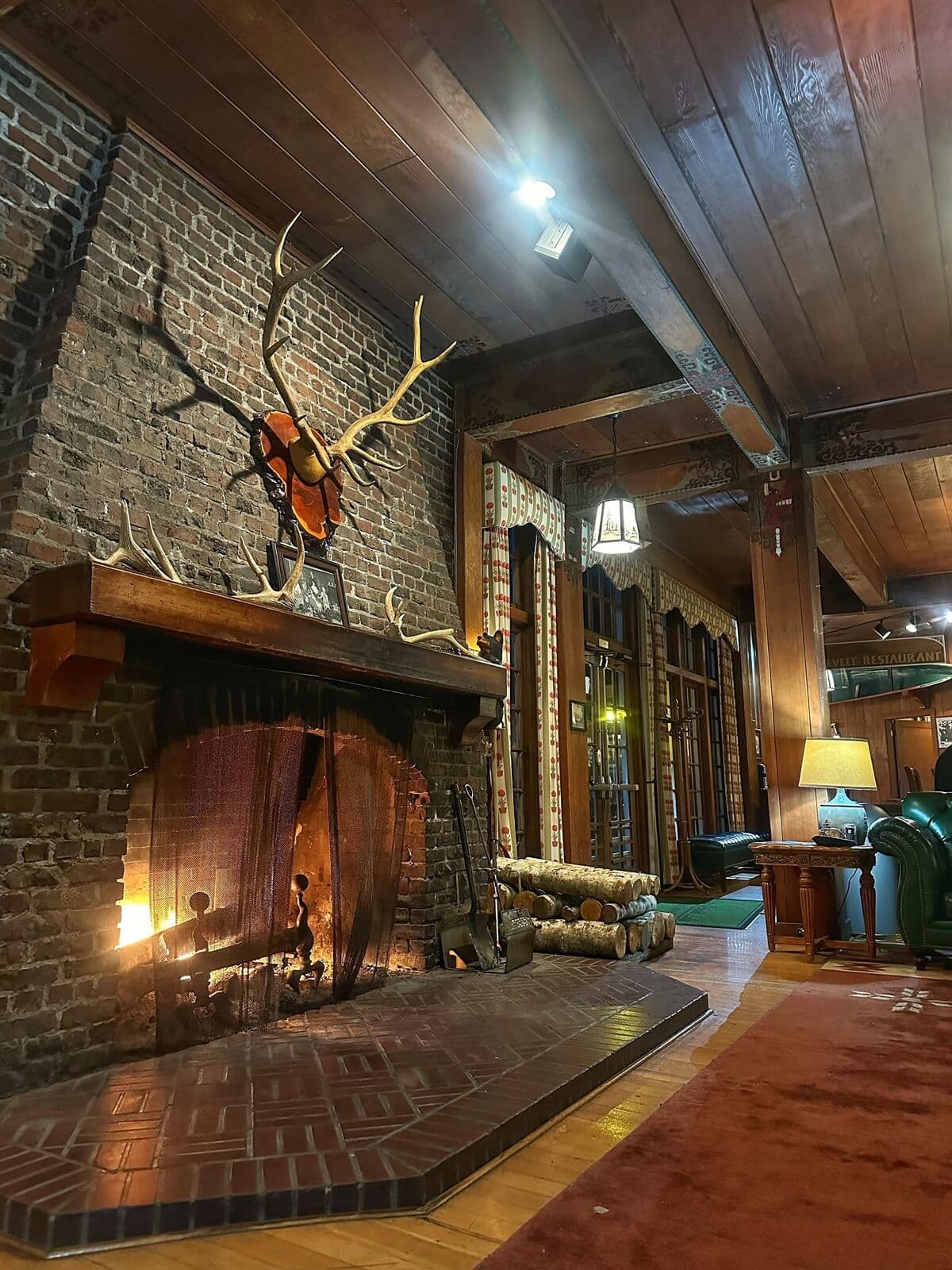 Cozy Lake Quinault Lodge interior with a roaring fire in a brick fireplace, topped by mounted antlers and rustic decor. Stacks of firewood, leather chairs, and warm wood-paneled walls create a welcoming atmosphere ideal for relaxing after a day outdoors.