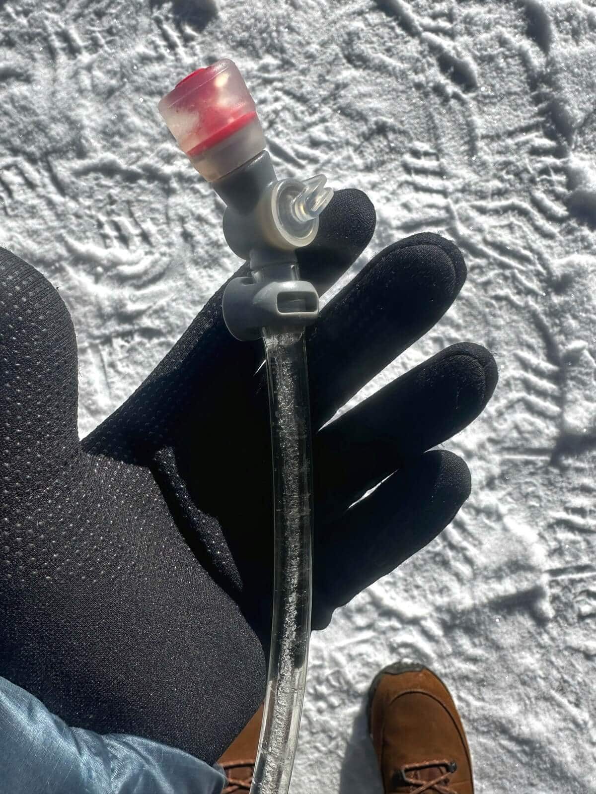 Close-up of a gloved hand holding a frozen hydration pack hose with visible ice crystals inside the tube, showing what it’s like to hike during winter in Olympic National Park. Snow-covered ground and brown winter boots are visible in the background.