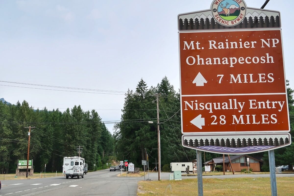 Roadside sign on the White Pass Scenic Byway shows directions to Mt. Rainier National Park, with Ohanapecosh 7 miles ahead and the Nisqually Entry 28 miles to the left. An RV drives toward a forested area under an overcast sky.