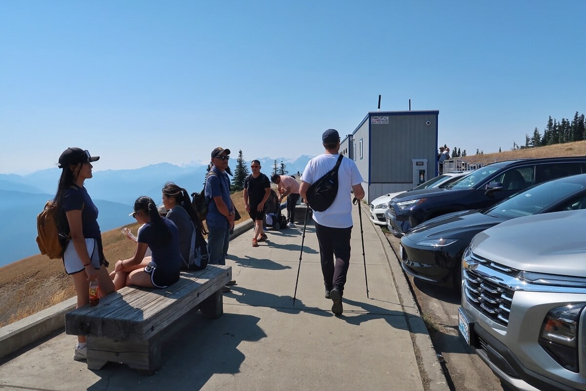 Image showing how to visit Hurricane Ridge, with hikers and visitors gathering near the temporary visitor center and parking area. The scene highlights accessible paved walkways, benches, and mountain views.