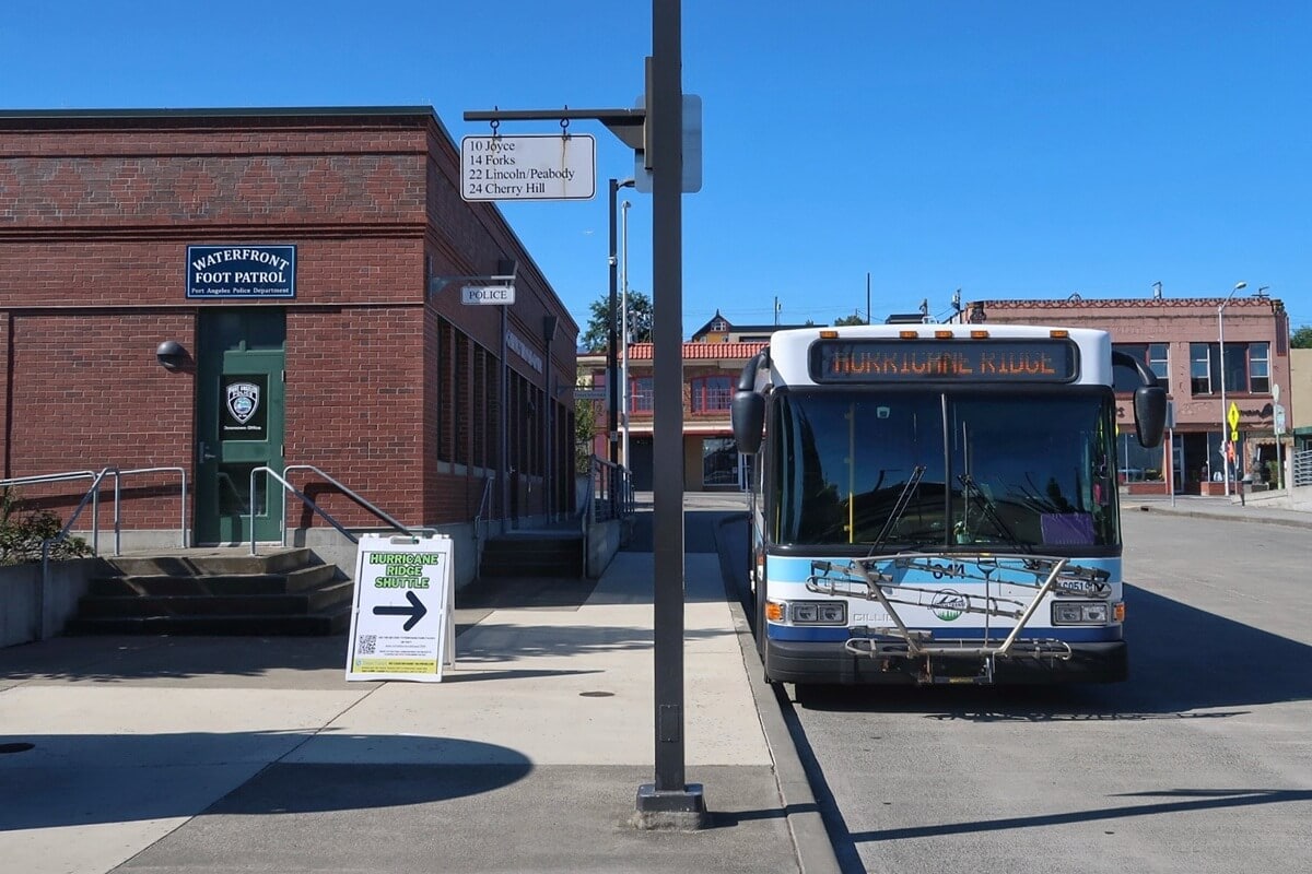 A shuttle bus heading to Hurricane Ridge from Port Angeles, parked outside a downtown building with a sidewalk sign showing the pickup point.