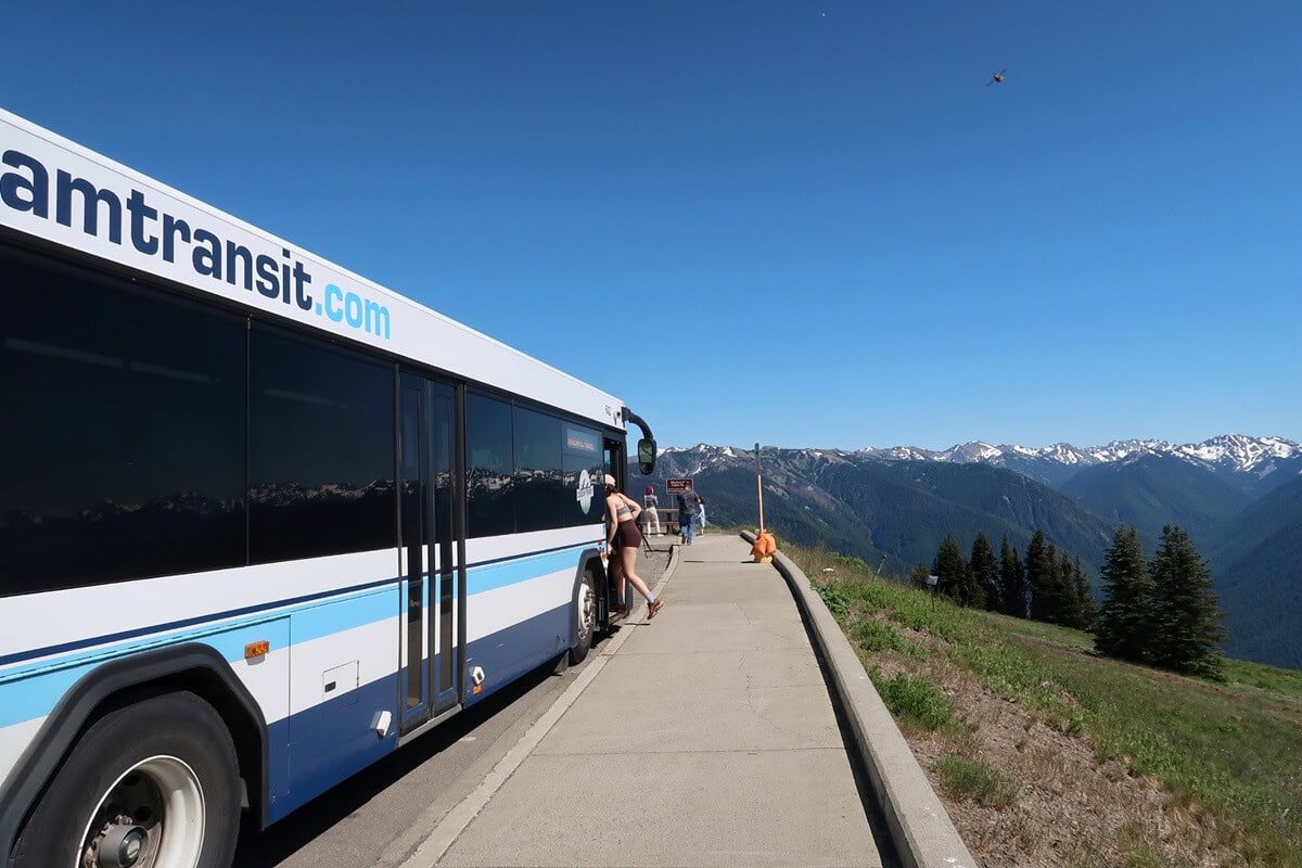 The Hurricane Ridge shuttle bus parked at a scenic overlook in Olympic National Park, with a passenger boarding and panoramic views of the snow-capped Olympic Mountains.