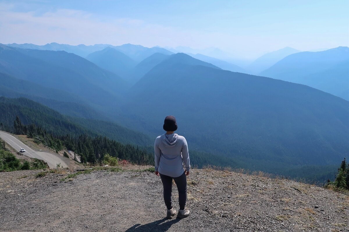 A hiker stands at a high vantage point taking in sweeping Hurricane Ridge views, with rolling blue-toned mountain ridges stretching endlessly into the hazy horizon with a winding road below. 