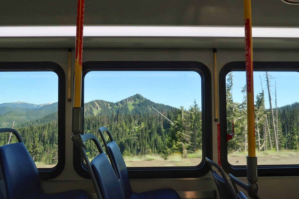 View from inside the Olympic National Park shuttle showing empty blue seats and large windows framing a scenic landscape of forested hills and a green, tree-covered mountain under a clear blue sky.