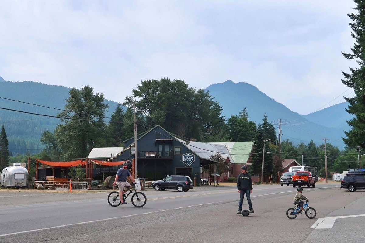 Three people in Packwood, one of the best towns near Mt Rainier, ride across a street with forested mountains in the background. The scene includes a brewery and vintage trailer, capturing the laid-back charm of mountain towns.