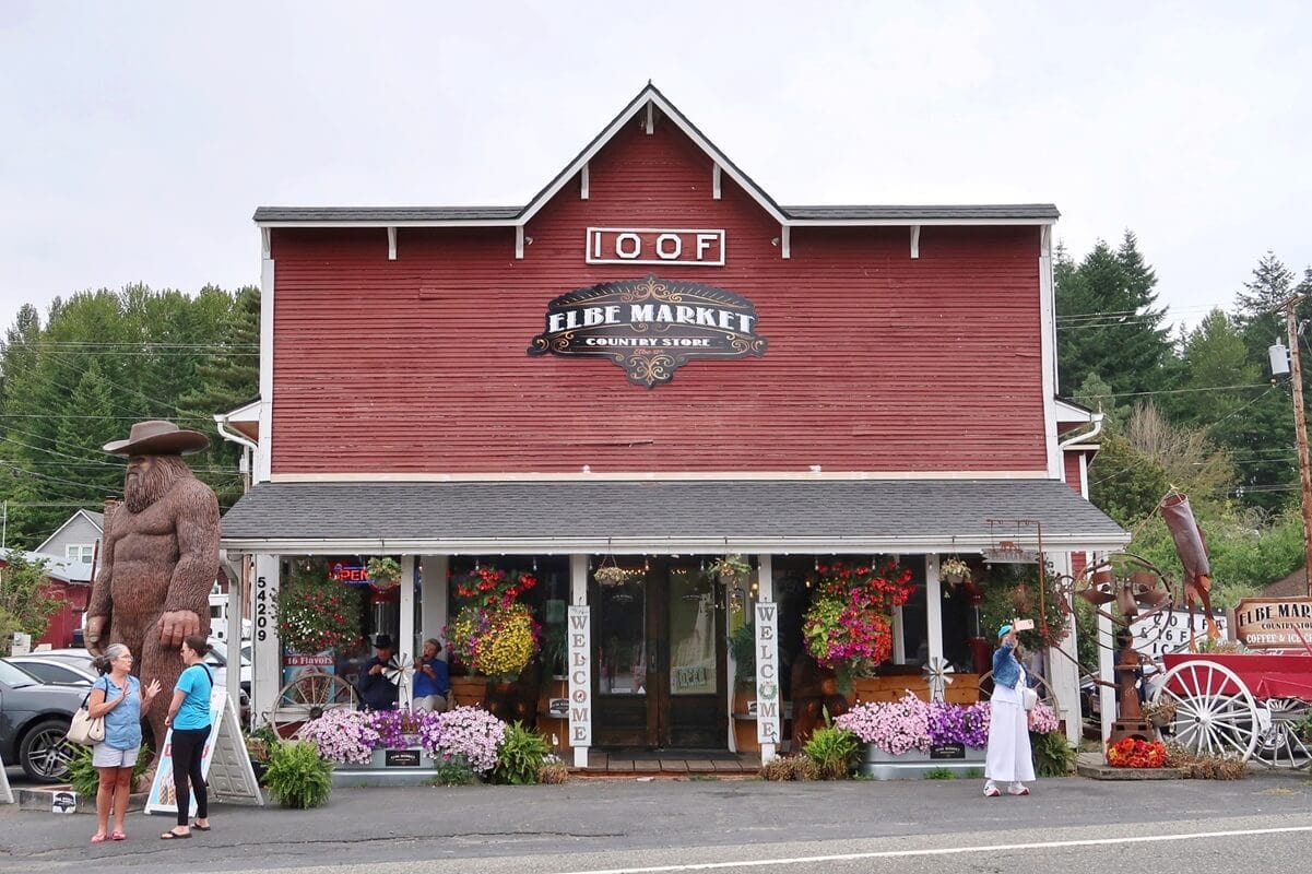Front view of the Elbe Market in Elbe, WA, housed in a rustic red building with white trim and a vintage sign. The storefront is decorated with colorful flowers, hanging baskets, and a large wooden Bigfoot statue wearing a cowboy hat, while visitors chat and take photos outside.