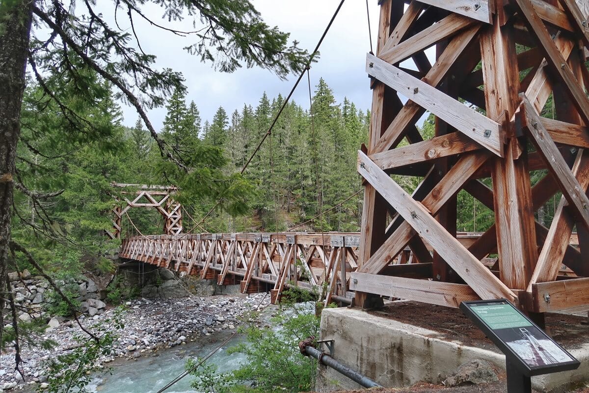 Close-up view of the Longmire Historic Suspension Bridge, a wooden truss-style structure spanning a rocky, glacial river with a dense evergreen forest in the background. The bridge’s massive timber supports and interpretive sign highlight its historic and architectural significance.