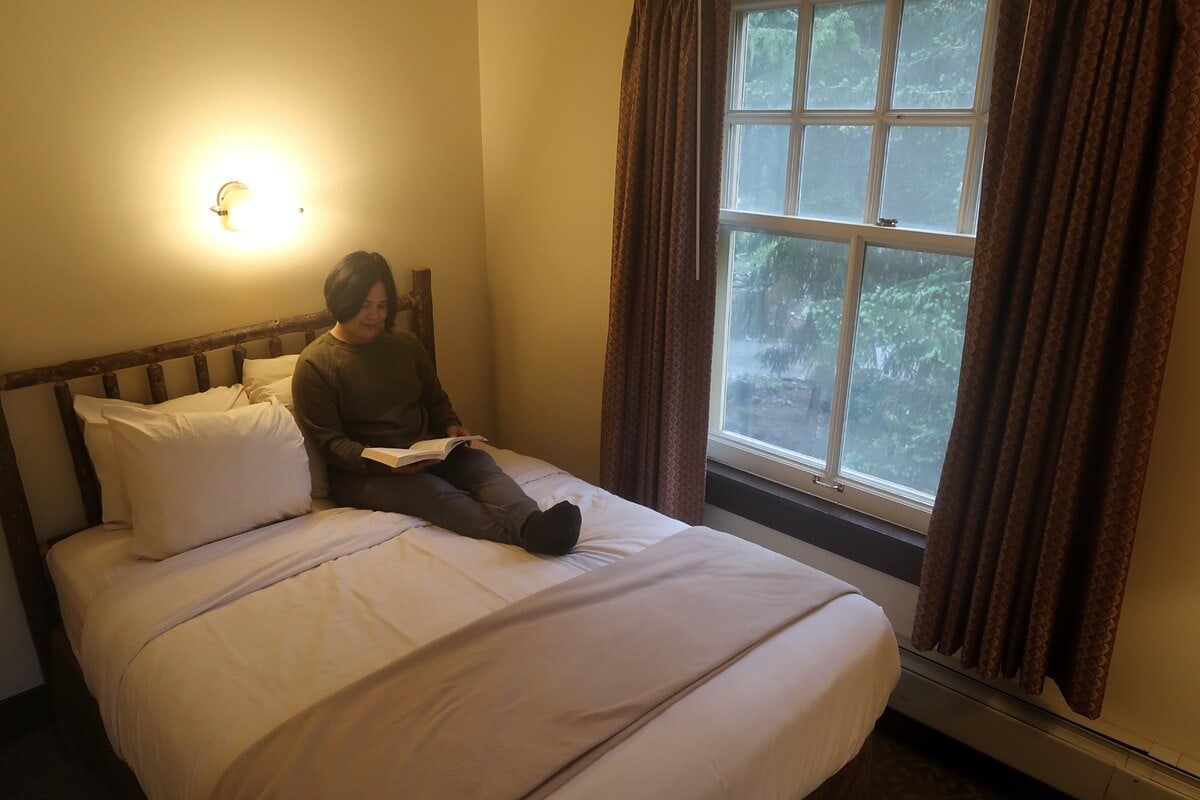 Inside a room at the National Park Inn Rainier, a woman reads a book while sitting on a neatly made bed with a rustic wood headboard. Soft lighting and a window view of evergreen trees create a calm, cozy atmosphere.
